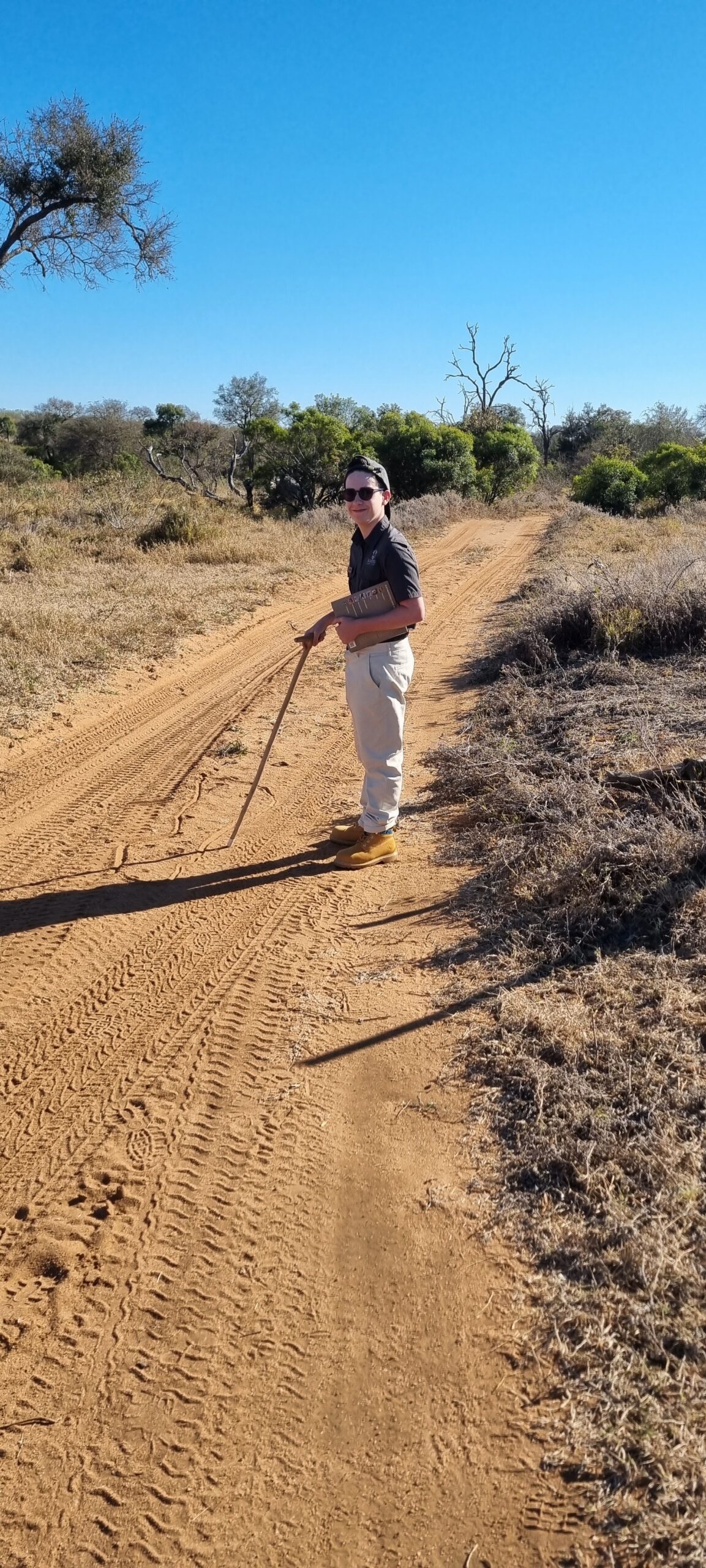 George standing on a sandy bush track holding a tracking stick and a tracks-and-signs guide